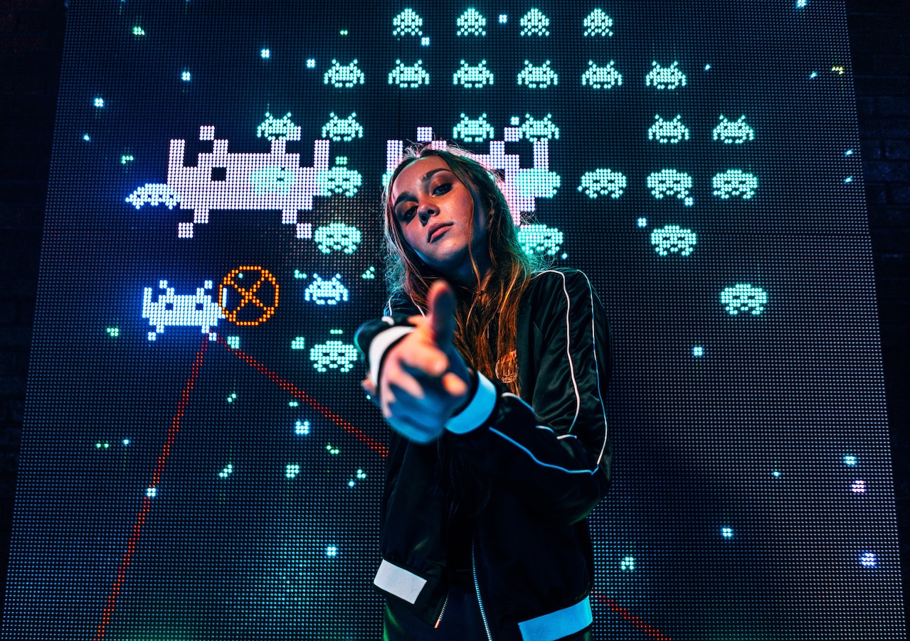 Writing challenges - Woman in jacket aiming finger guns at the camera, with Space Invaders game background
